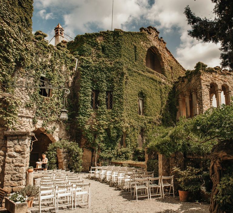 Rustic outdoor wedding ceremony at La Baronia Barcelona complete with greenery across castle and white chairs lining the aisle