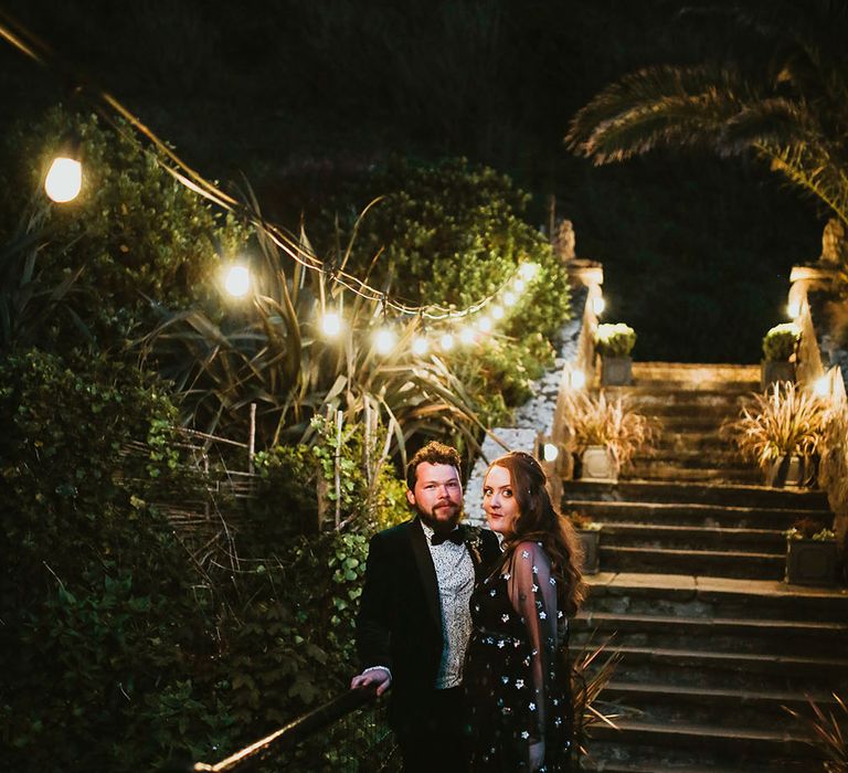 Bride wearing black celestial reception dress and groom in velvet dinner jacket stand beneath festoon lighting outdoors 