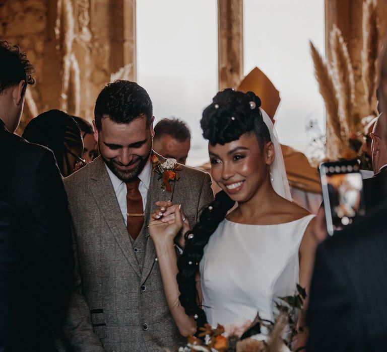 Bride wears her black hair in intricate braided style with pearl hair accessories as she leaves Christian ceremony with her groom in three piece suit