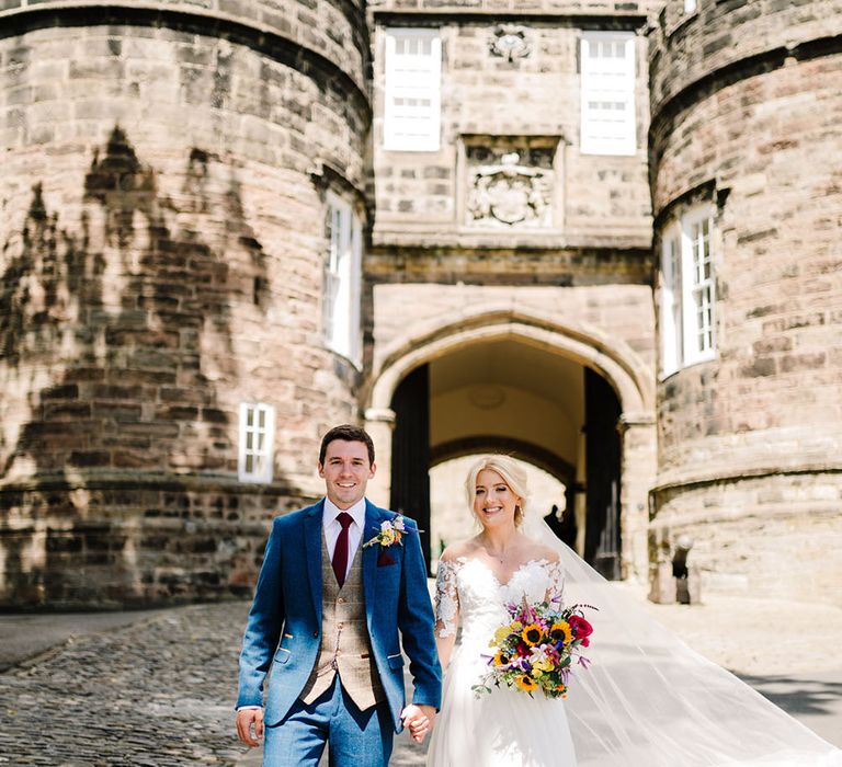 Bride holds bright floral bouquet with sunflowers and walks hand in hand with her groom wearing three piece suit 