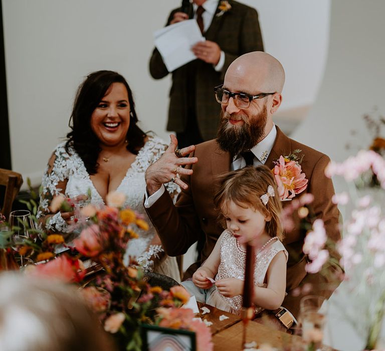 Bride & groom sit with their daughter during industrial styled wedding reception complete with orange fabric table runners and wooden banquet tables