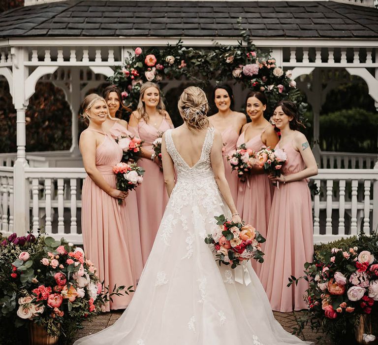 Bridesmaids in pink dresses in front of a pink flower arch holding pink bouquets with the bride showing off her elegant updo 