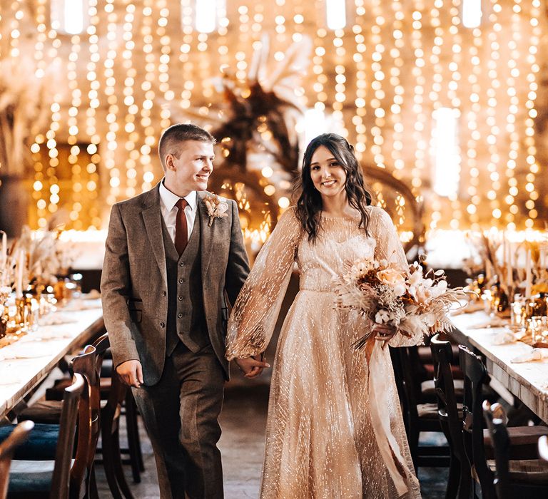 Bride & groom walk through wooden banquet tables as fairy light canopy shines in the background