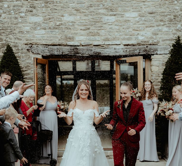 Bride in a burgundy suit jacket and trousers with a black shirt having a confetti moment with bride in a strapless flower wedding dress