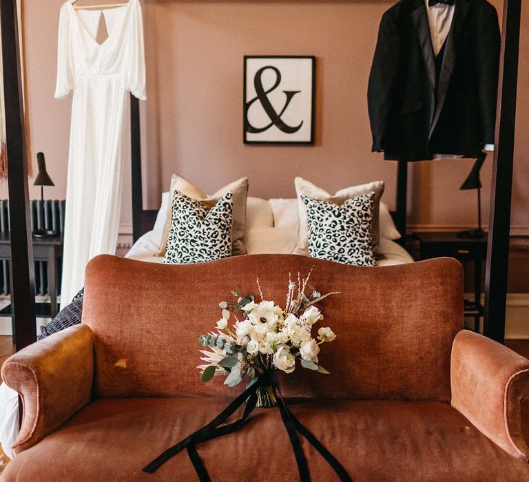 Bedroom at Pepper Arden Hall with bridal and groom outfits hanging, leopard print pillows, crushed orange chaise lounge and minimalistic, neutral floral bouquet