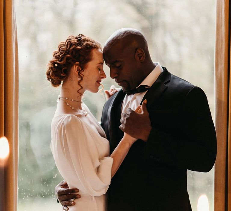 Bride in satin quarter length sleeve wedding dress and groom in black tux embracing and dancing at candlelight 