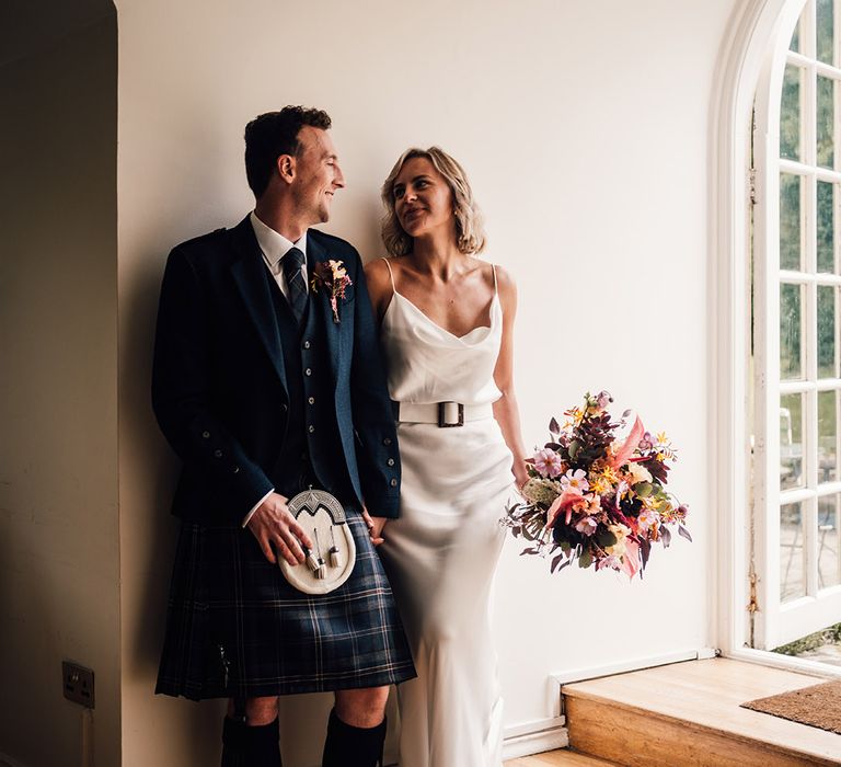 Bride in silk wedding dress looks lovingly toward her groom in kilt after wedding ceremony