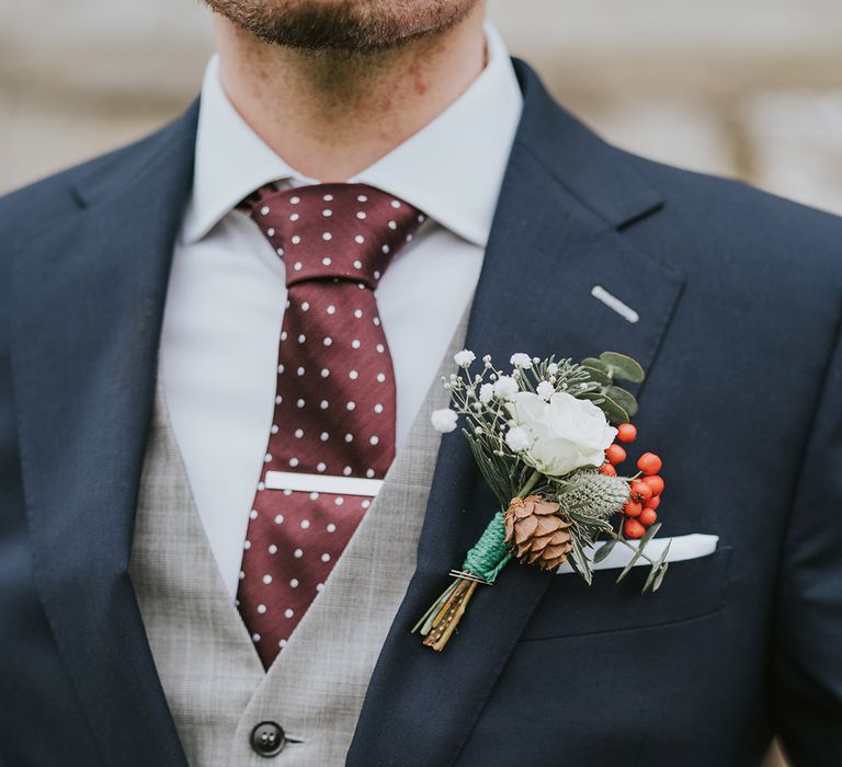 Groom in navy blue suit jacket, light grey waistcoat and burgundy tie with white spots and winter buttonhole 