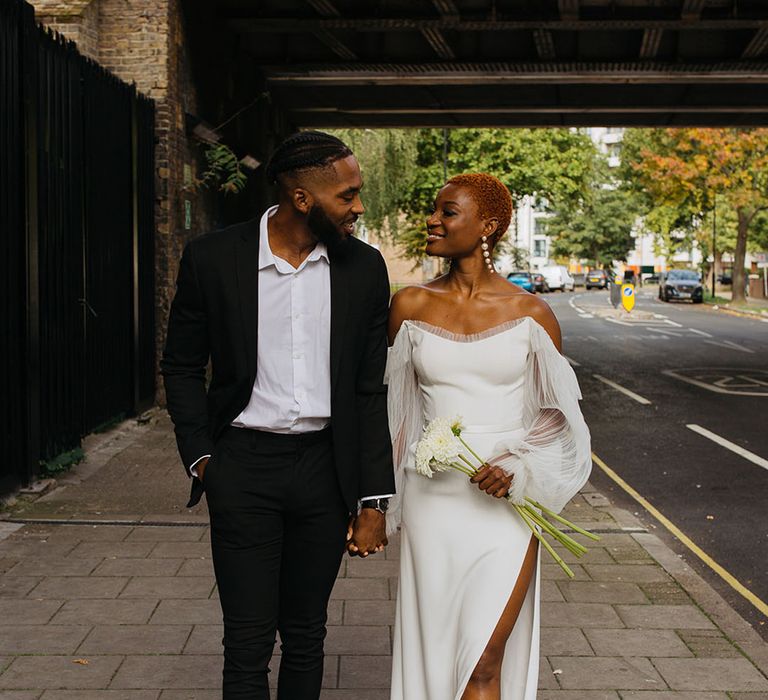 Bride in off shoulder wedding dress with lace puff sleeves and leg slit holding hands with groom in skinny black suit