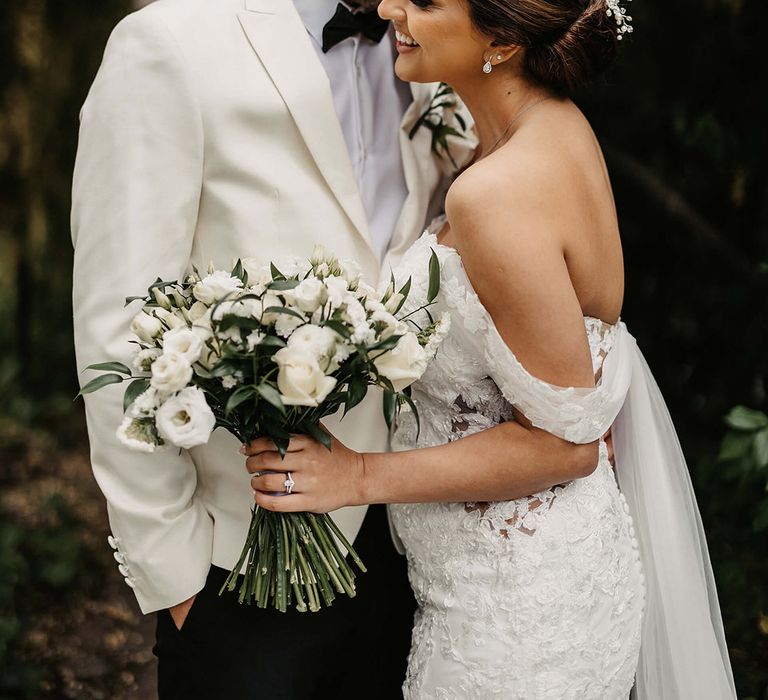 The groom in a white suit jacket embraces the bride in an off the shoulder flower wedding dress holding a white flower bouquet 