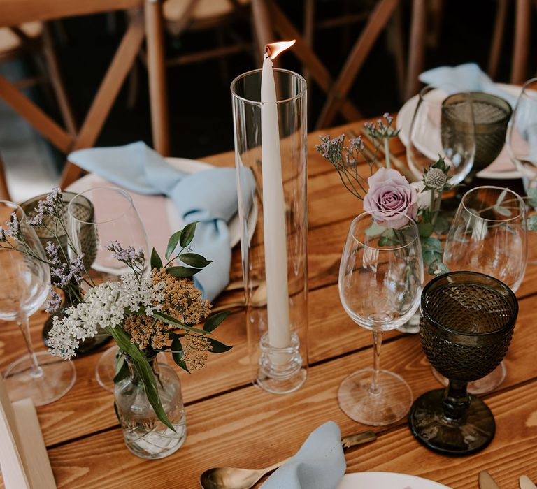 White taper candle in glass vase with white plates, blue napkins and pink wedding menus and gold cutlery