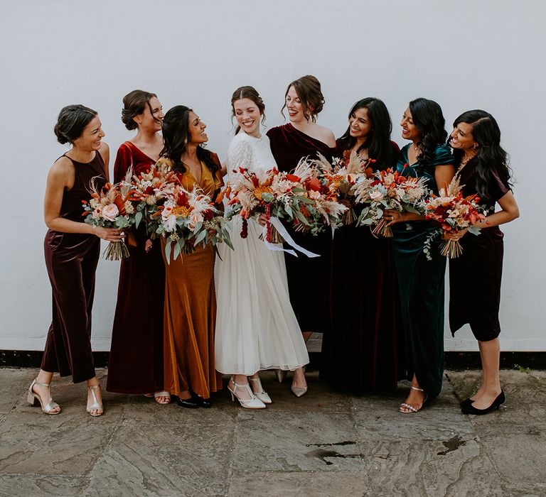 Bride in long sleeve lace vintage wedding dress and closed toe satin heels standing with bridesmaids in mismatched velvet bridesmaid dresses all holding large autumnal bouquets with orange, black, green and yellow roses, peonies, foliage and pampas grass