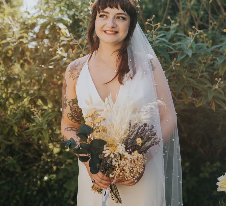 Bride in silk-look v-neck sleeveless ASOS wedding dress and gold halo headband with sparkly stars and pearl detailed veil holding dried flower wedding bouquet with white bunny tails, green foliage, yellow dried gypsophila, lavender and baby’s-breath with white ribbon