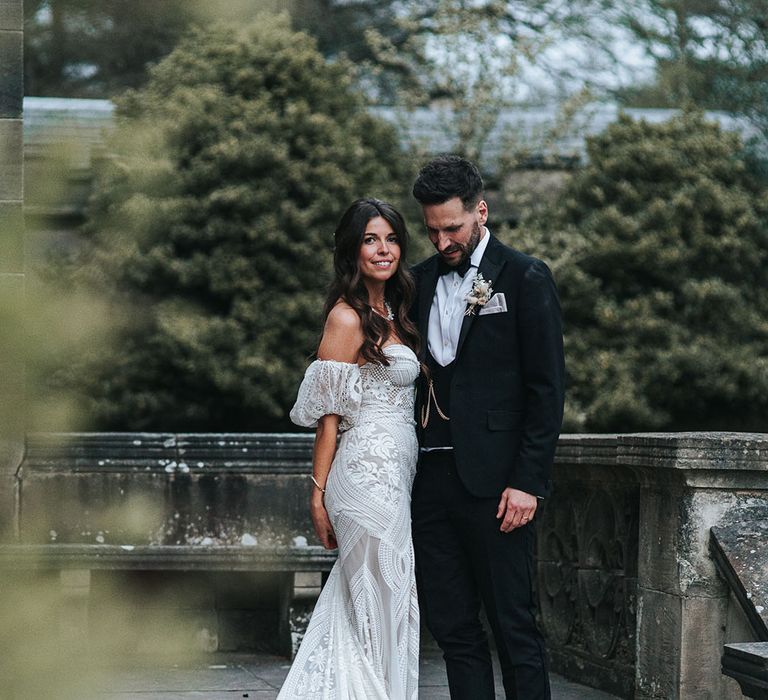 Bride with brown hair in a half up half down style wearing a strapless fitted boho wedding dress standing with the groom in black tie 