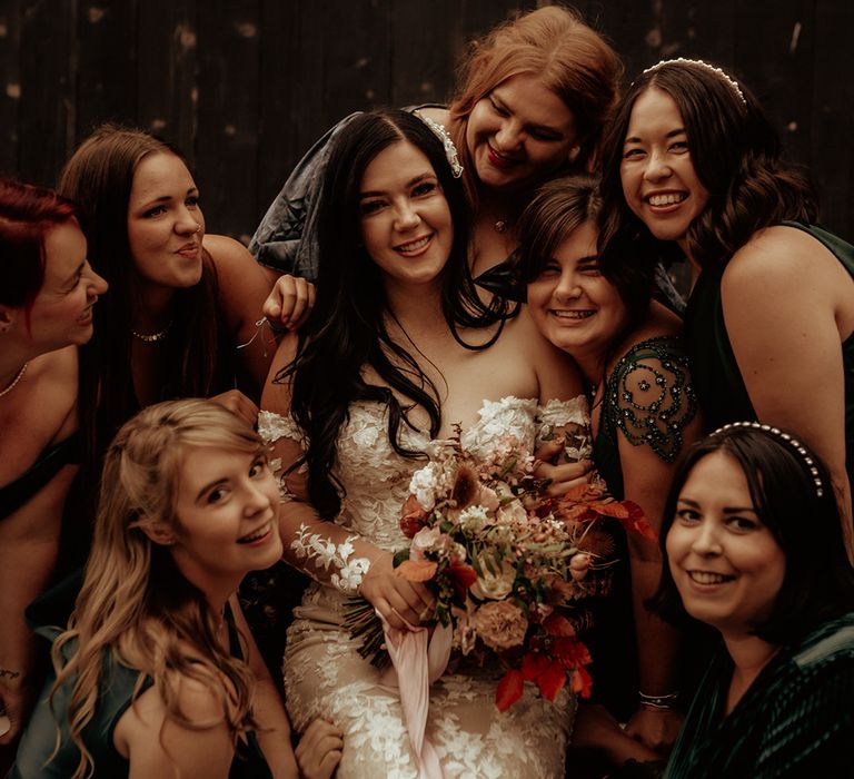 Bride sitting front and centre wearing off the shoulder Abigail Westrup bridal gown with long, sheer sleeve and lace detailing holding dried flower bouquet with roses, eucalyptus, peonies and autumn leaves with bridal party standing around her in dark green mismatched bridesmaid dresses 