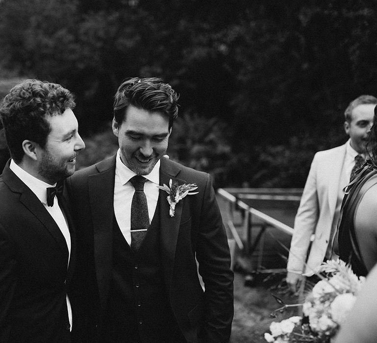 Groom wears three piece suit complete with floral buttonhole and tie pin in black and white image 