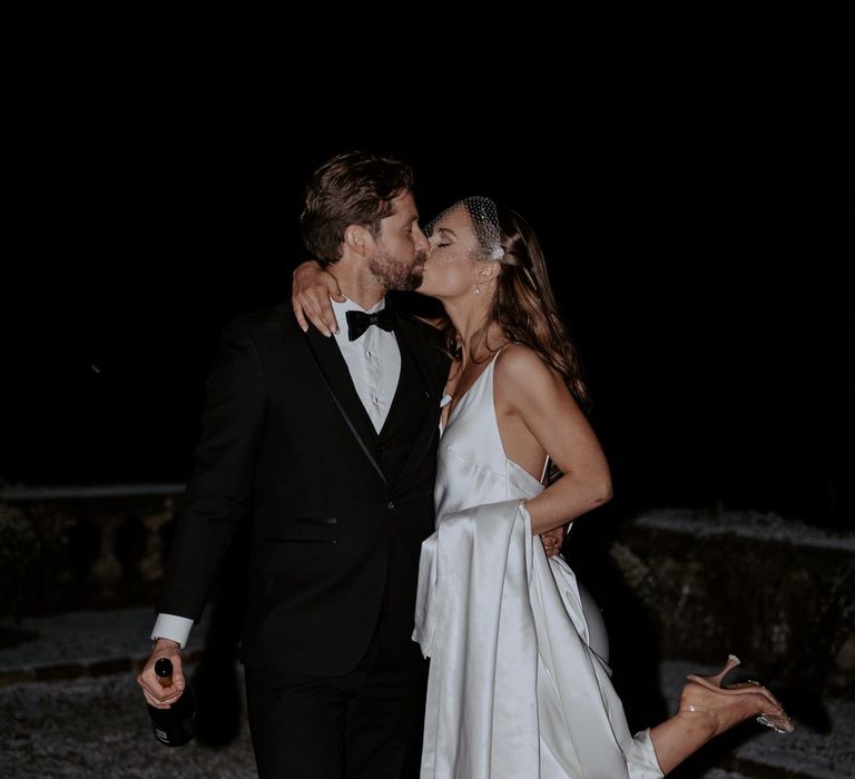 Bride in satin cowl neck wedding dress with open-toe clear heels and groom in classic black tux kissing on the grounds of Eaves Hall