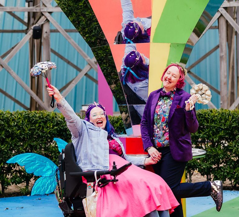 Bride in wheelchair with blue fairy wings wearing personalised denim jacket, pink wedding dress and blue fishnet tights holding hands with bride in purple velvet blazer and black suit trousers with purple bowtie, pink patterned pocket square and colourful patterned shirt on rainbow floor 