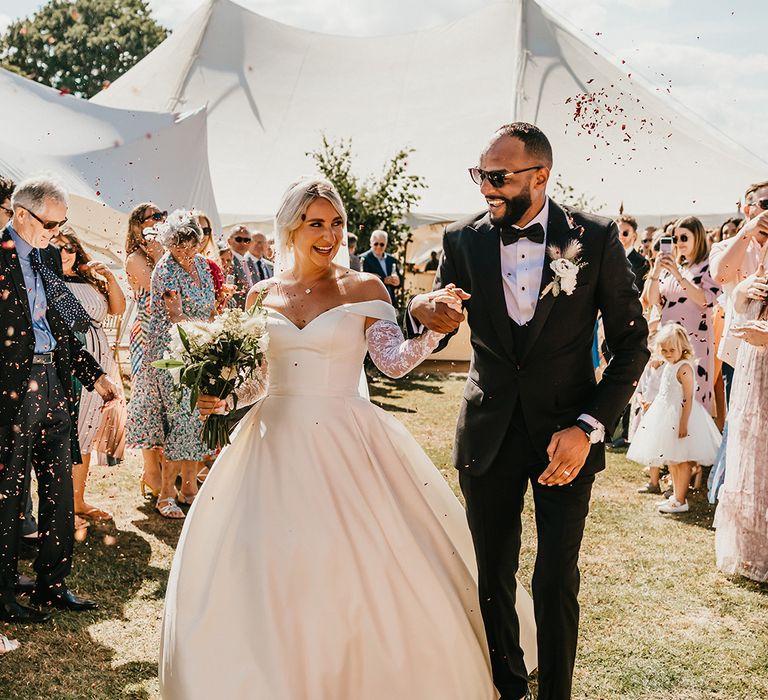 Bride in off-the-shoulder princess wedding dress walks through colourful confetti exit alongside her groom