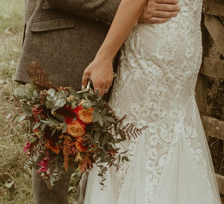 Bride in fitted lace off the shoulder boho wedding dress holding orange autumnal bouquet with the groom in a three piece tweed suit 