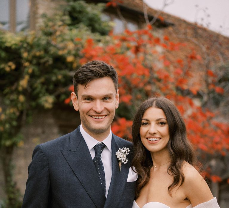 Groom in dark navy suit with spotted tie and white flower buttonhole with the bride in a strapless wedding dress with detachable wedding sleeves with pearls 