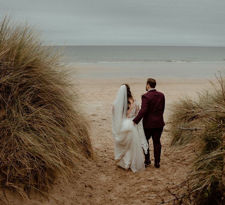 The bride in a boho lace wedding dress walking with the groom in a burgundy suit along the coast 