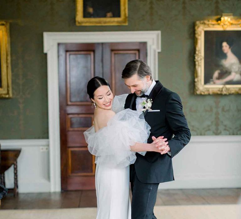 Bride in strapless wedding dress with puff tulle sleeves dancing with groom in classic black tux 