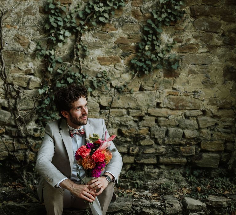 Groom in greige suit and colourful bow tie sitting down with bridal bouquet consisting of red pincushion flowers, coral carnations, magenta peonies and anthuriums