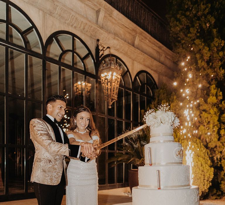 Bride and groom cutting four tier buttercream wedding cake with mini sparkler decorations, white flower wedding cake topper and intricate detailing with large golden sword