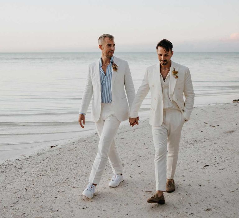 Groom wearing white blazer, light blue and white striped shirt, dusky orange garden rose and dried flower boutonniere holding hands with groom in mens white wedding suit with dusky orange garden rose and dried flower boutonniere, white shirt and silver jewellery 