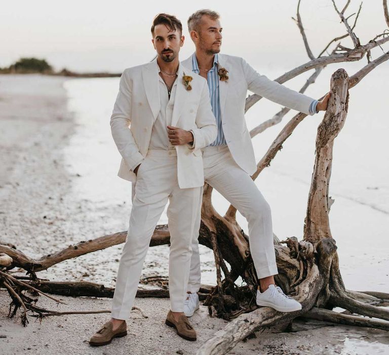 Groom in mens white wedding suit with dusky orange garden rose and dried flower boutonniere, white shirt and silver jewellery posing with groom in white blazer, light blue and white striped shirt, dusky orange garden rose and dried flower boutonniere and gold jewellery posing by some driftwood 