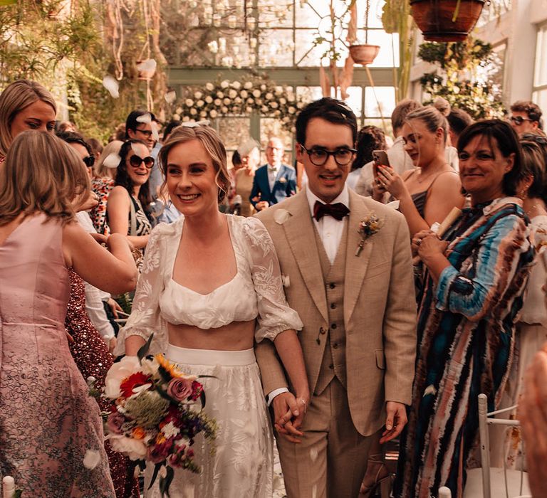 Bride in lace bridal separates walking back down the aisle with the groom in a cream suit as a married couple to white petal confetti 