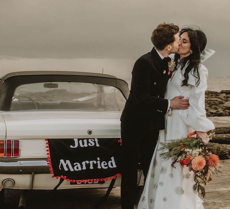 The groom in a black suit kisses the bride in a wedding jumpsuit with sparkly detail in front of wedding car with wedding banner 