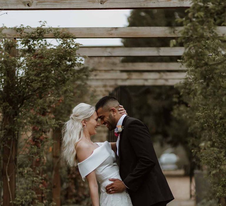 Groom in classic three piece grooms tuxedo with colourful spring boutonniere embracing bride in off the shoulder lace wedding dress holding colourful spring bridal bouquet 