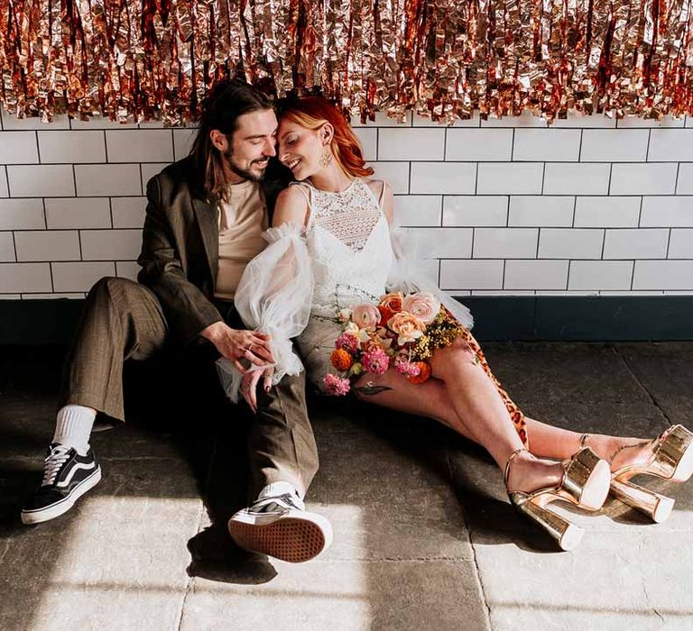 Groom in brown blazer, white shirt, grey suit trousers and vans shoes sitting on the floor of Northern Monk Refectory Leeds with bride in 70s-inspired boatneck short lace wedding dress with detachable tulle puff sleeves and platform gold wedding heels 