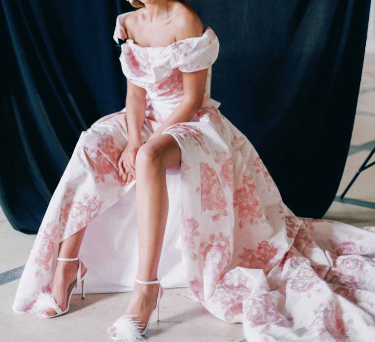 Bride in off the shoulder white and pink floral patterned bridal gown with black bow and front slit sitting on chair at Settrington Orangery bridal shoot