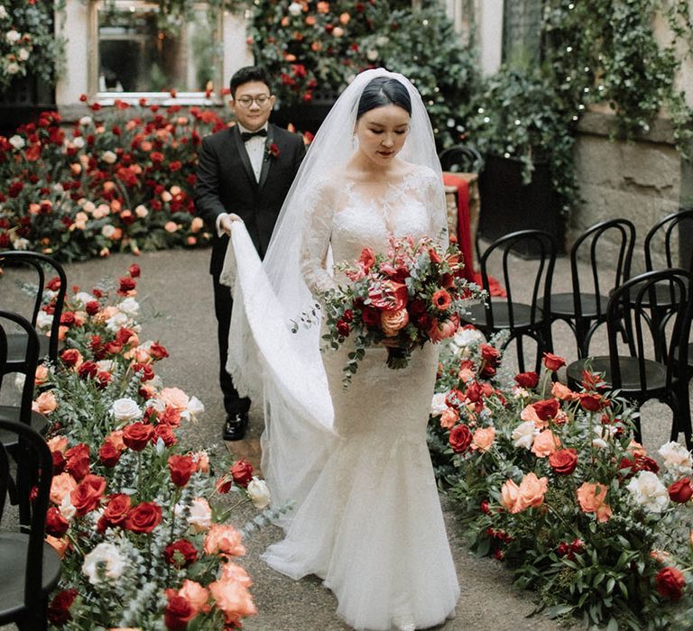 The groom in a black tuxedo walks back down the aisle with the bride carrying her train who wears a mermaid lace wedding dress 