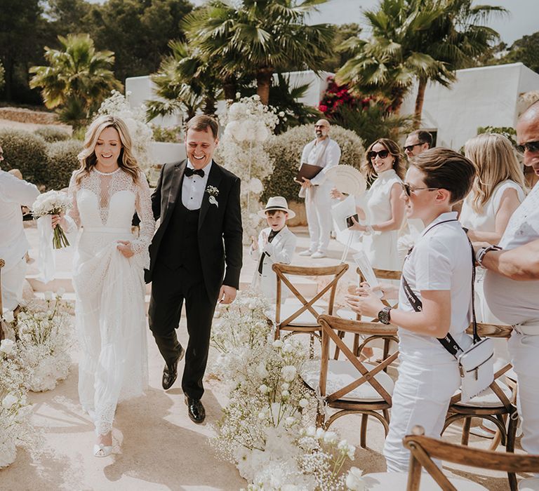 bride in a lace wedding dress and groom in a tuxedo descending up the aisle of white floor flowers as husband and wife