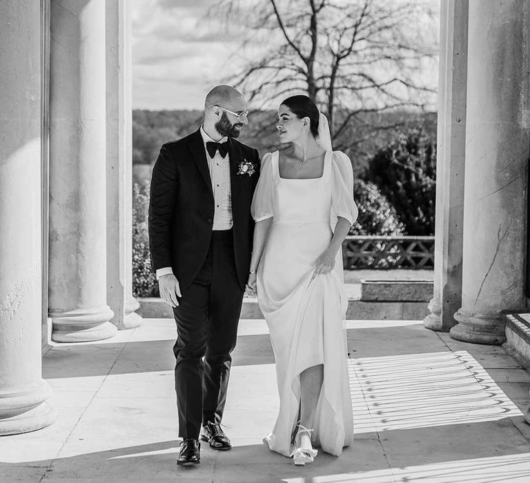 Bride in short sheer puff sleeve wedding dress with square neck and wrist loop with sheer pearl embellished bridal veil walking with groom in classic black tuxedo with bowtie and wildflower boutonniere at Buxted Park