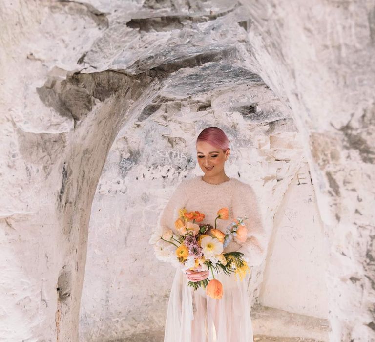 Bride in sheer overlay wedding dress and fluffy bridal coverup holding orange bridal bouquet in Margate Caves 