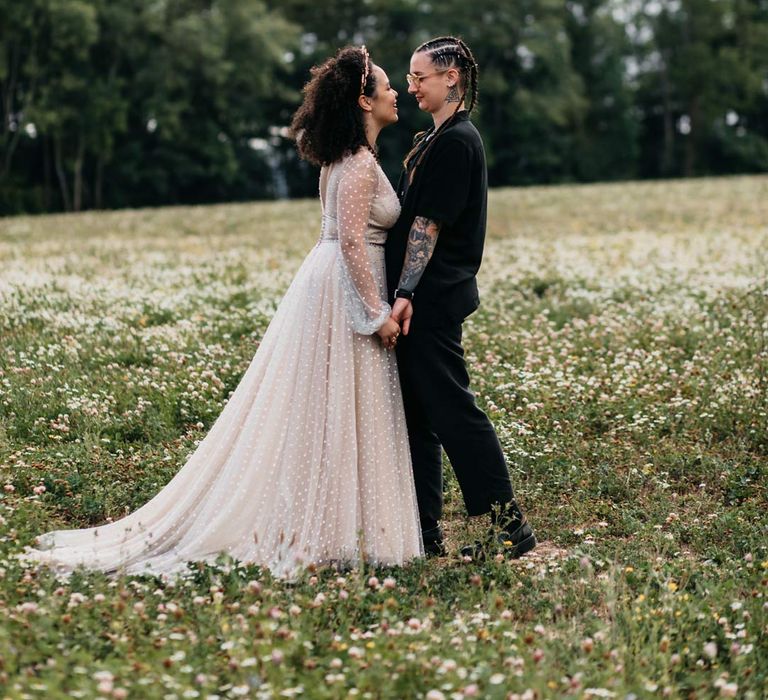 Bride in silver chains, glasses, black shirt and black trousers holding hands with bride in long sleeve v-neck sheer polka dot overlay sparkly wedding dress and gold celestial bridal headband 