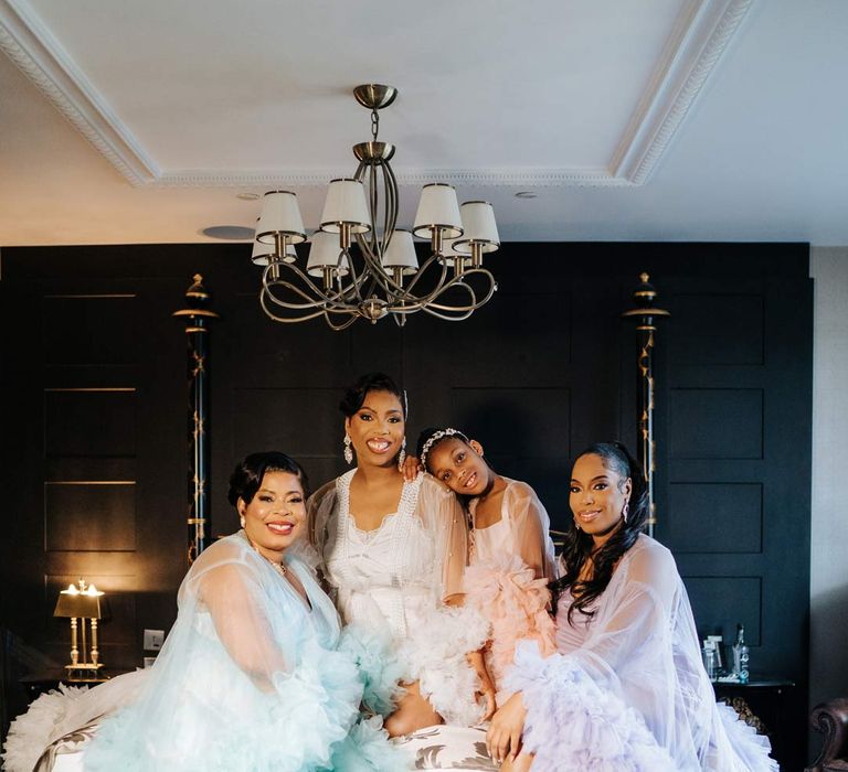 Bride in white sheer tulle and pearl bridal getting ready robe with bridesmaids and flower girl in mismatched pastel sheer tulle and pearl bridal getting ready robes sitting on bed at Syon Park wedding venue