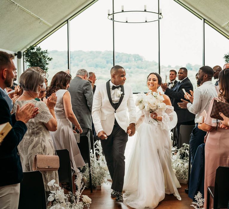 Bride and groom walking up the aisle at crumplebury wedding wearing a white off-the-shoulder wedding dress and white tuxedo