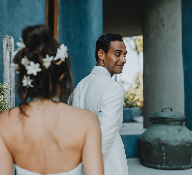 Bride in strapless wedding dress with flower hair accessories approaches groom in cream groom suit during first look photos