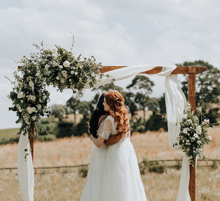 Brides embrace each other to celebrate the wedding with the wooden altar decoration covered in white flowers 