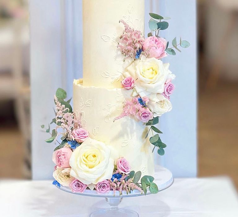 Three tier white frosted wedding cake with white rose wedding flowers and stencilled details 