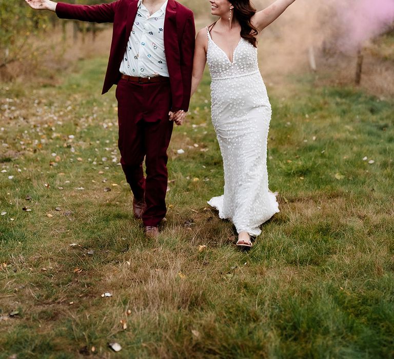 Groom in burgundy suit and bride in lace wedding dress walk together as they wave orange and pink smoke bombs 