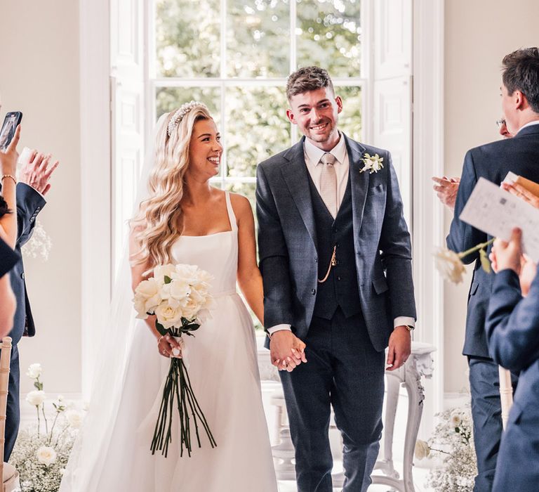 bride-carrying-white-rose-bouquet-down-the-aisle-with-groom-in-navy-suit