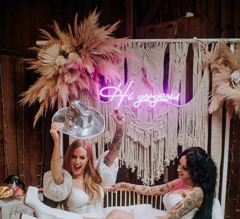 bride-wearing-retro-white-platform-heeled-boots-with-macarame-photo-backdrop-smiling-at-bride-with-disco-cowboy-hat