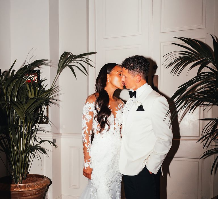 bride-in-illusion-lace-long-sleeve-wedding-dress-kissing-groom-in-white-tuxedo